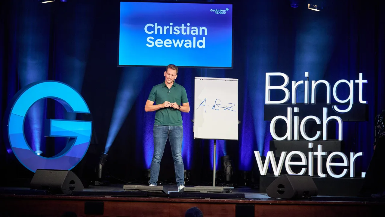 Christian Seewald auf der Bühne der GEDANKENtanken-Rednernacht, vor einem Flipchart, hinter ihm die Leuchtschrift 'Bringt dich weiter'